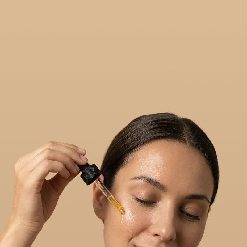 Woman applying a dropper of oil to her face with a beige background